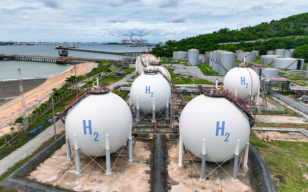 Industrial hydrogen storage tanks (H2) at a coastal energy facility with pipelines, large spherical containers, and port infrastructure in the background